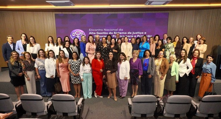 A imagem mostra um grande grupo de mulheres e alguns homens reunidos em um auditório para um eve...